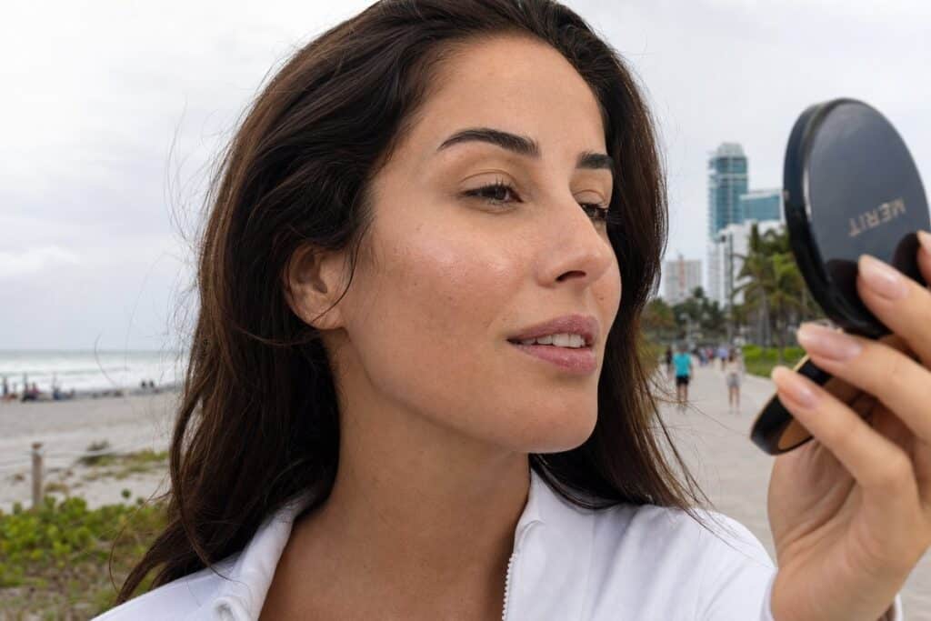 Woman admiring her glowing post-chemical peel skin in a compact mirror on a Miami beach boardwalk