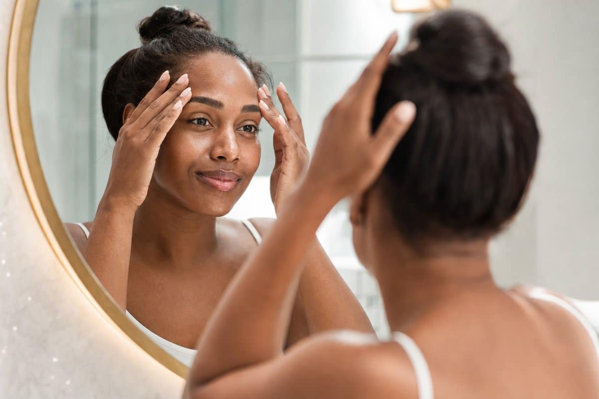 Black woman in her 30s viewing her smooth, even-toned cheeks after a VI Peel in a bathroom mirror