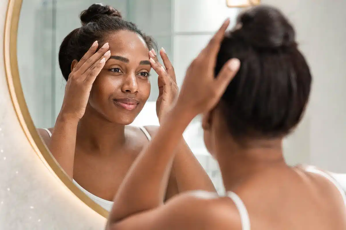 Black woman in her 30s viewing her smooth, even-toned cheeks after a VI Peel in a bathroom mirror