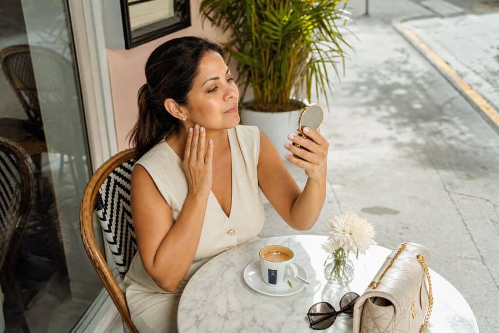 Latina woman in her 40s admiring her smooth, even skin texture in a compact mirror at a Miami café after a skin resurfacing treatment