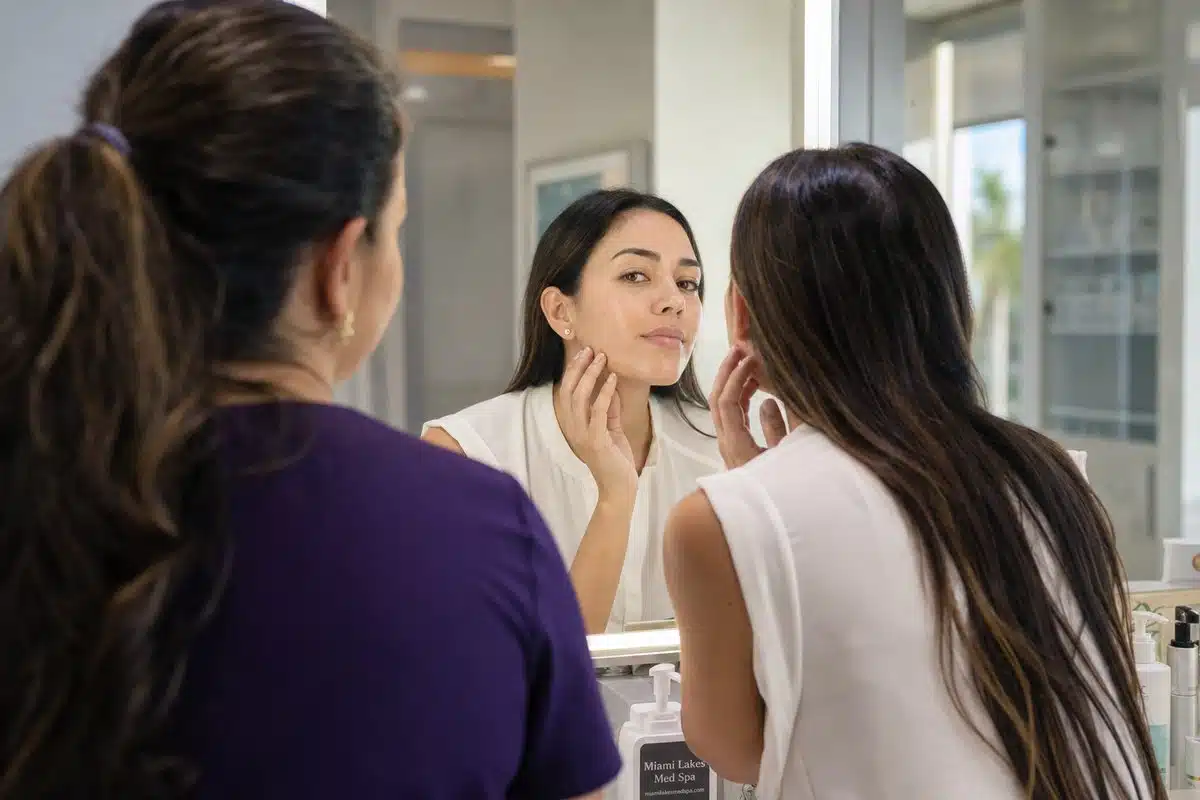 Woman in her late 20s at a vanity mirror showing smooth, spot-free cheek skin after microneedling and chemical peel treatment