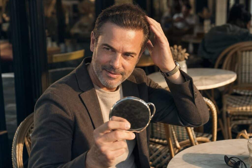 Man in his late 40s at a café checking his restored hairline in a compact mirror after hair restoration treatment