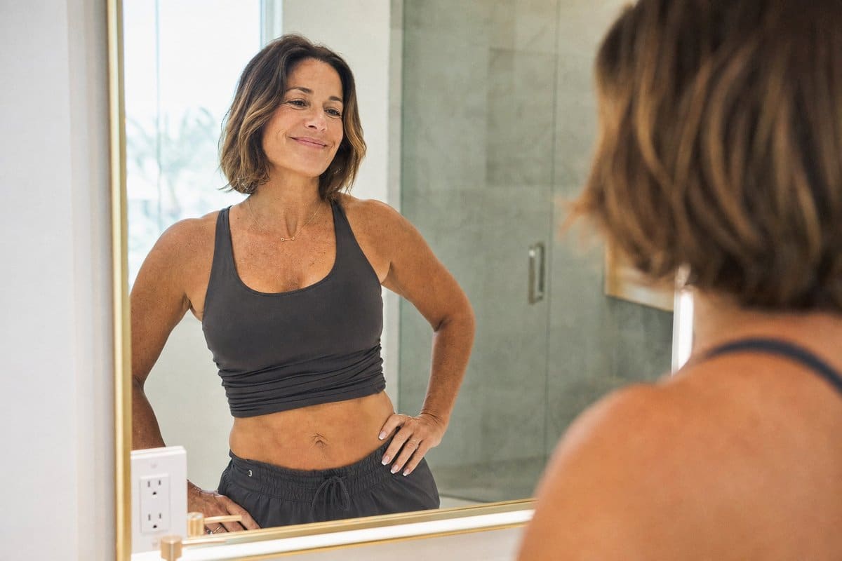 Woman in her 50s admiring her slimmer waistline in a mirror after breaking through a GLP-1 weight loss plateau